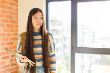 Genç Asyalı kadın şaşkın ve şaşkın görünüyor, merak ediyor ya da bir sorunu çözmeye çalışıyor veya düşünüyor.