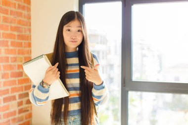 Genç Asyalı kadın, şaşkın ve şaşkın bir bakışla kendini işaret ediyor. Seçildiğine şaşırmış ve şaşırmış.