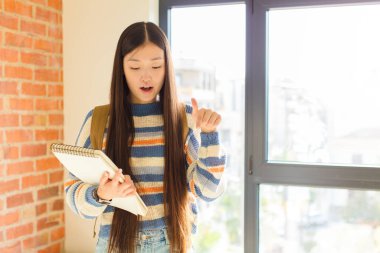 Genç Asyalı kadın şok olmuş, ağzı açık ve şaşırmış hissediyor. İnançsızlık ve şaşkınlık içinde bakıyor ve işaret ediyor.