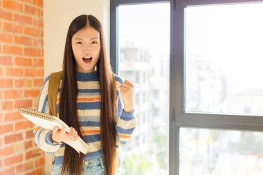 Asyalı genç bir kadın öfkeli bir ifadeyle bağırıyor ya da yumruklarıyla başarıyı kutluyor.