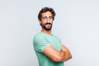 Genç sakallı adam kameraya çapraz kollarıyla gülümsüyor ve mutlu, kendinden emin, tatmin olmuş bir yüz ifadesi var.