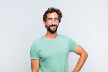 Genç sakallı adam mutlu bir şekilde gülümsüyor, eli kalçasında ve kendinden emin, olumlu, gururlu ve arkadaş canlısı.