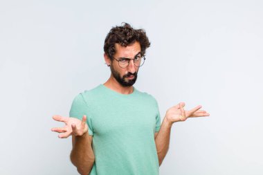 Genç sakallı adam şaşkın, kafası karışmış ve stresli görünüyor, farklı seçenekler arasında merak ediyor, kararsız hissediyor.