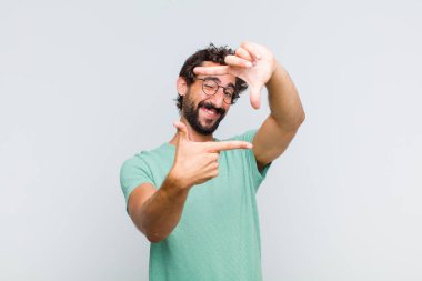 Genç sakallı adam mutlu, arkadaş canlısı ve pozitif hissediyor, gülümsüyor ve elleriyle bir portre veya fotoğraf çerçevesi yapıyor.