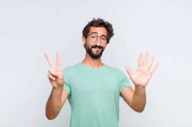 Genç sakallı adam gülümsüyor ve dost canlısı görünüyor, 7 ya da 7 numarayı gösteriyor, eli önde, geri sayıyor.