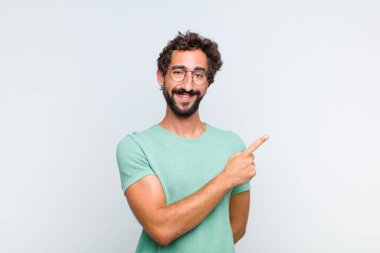 Genç sakallı adam neşeyle gülümsüyor, mutlu hissediyor ve yan ve yukarı doğru işaret ediyor, fotokopi uzayındaki nesneleri gösteriyor.