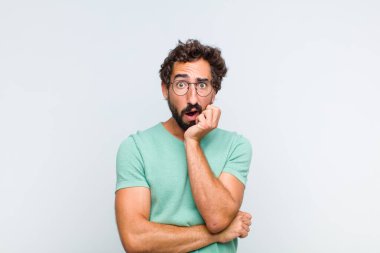 Genç sakallı adam şok ve inançsızlıkla ağzı açık, elleri yanağında ve kolları çaprazlanmış, sersemlemiş ve şaşırmış hissediyor