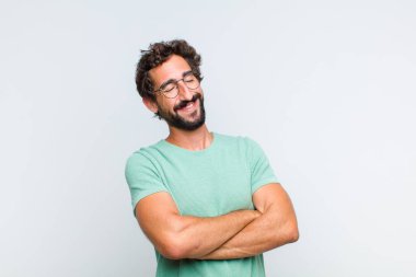Genç sakallı adam kollarını kavuşturup mutlu bir şekilde gülüyor, rahat, pozitif ve tatmin edici bir pozla.