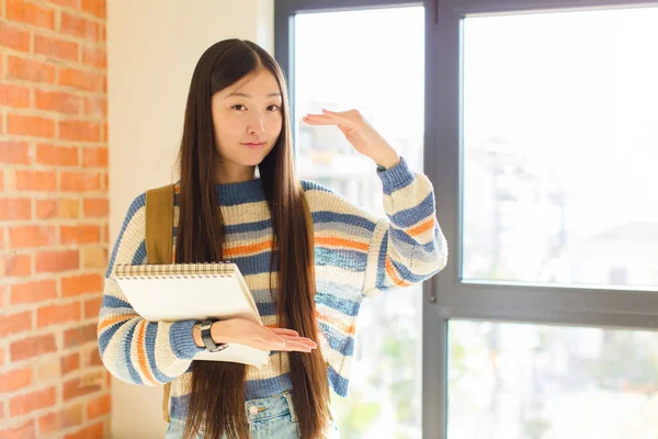 Genç Asyalı bir kadın iki eli de yan tarafında fotokopi alanı olan bir nesneyi tutuyor, bir nesneyi gösteriyor, sunuyor ya da reklamını yapıyor.