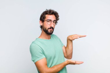 Genç sakallı adam bir objeyi iki eliyle fotokopi alanında tutuyor, bir nesneyi gösteriyor, sunuyor ya da reklamını yapıyor.