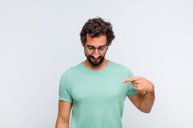 Genç sakallı adam neşeyle gülümsüyor, yere bakıyor ve göğsüne işaret ediyor.