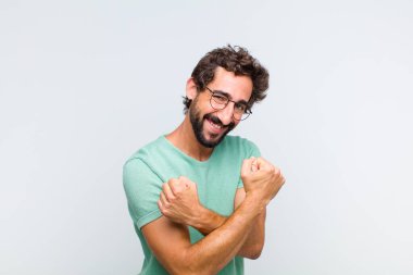 Genç sakallı adam sevinçle gülümsüyor ve kutluyor, yumruklarını sıkıp kollarını kavuşturuyor, mutlu ve pozitif hissediyor.