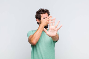 Genç sakallı bir adam yüzünü eliyle kapatıyor ve kamerayı durdurmak için diğer elini öne koyuyor. Fotoğraf ya da resimleri reddediyor.