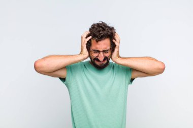 Genç sakallı adam stresli ve hayal kırıklığına uğramış hissediyor, ellerinizi havaya kaldırıyor, yorgun, mutsuz ve migren ağrısı çekiyor.