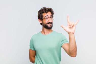 Genç sakallı adam mutlu, eğlenceli, kendinden emin, pozitif ve asi hissediyor, elle kaya ya da heavy metal işareti yapıyor.