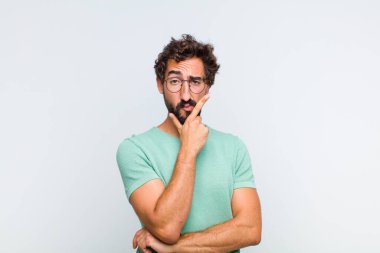 Genç sakallı adam ciddi, düşünceli ve güvensiz görünüyor, bir kolu çapraz ve çenesi üzerinde, seçenekleri tartıyor.