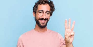 Genç sakallı adam gülümsüyor ve arkadaş canlısı görünüyor. Üç ya da üç numarayı gösteriyor. Elleri önde, geriye doğru sayıyor.