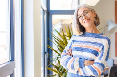 Orta yaşlı bir kadın kollarını kavuşturup mutlu bir şekilde gülüyor, rahat, pozitif ve tatmin edici bir pozla.
