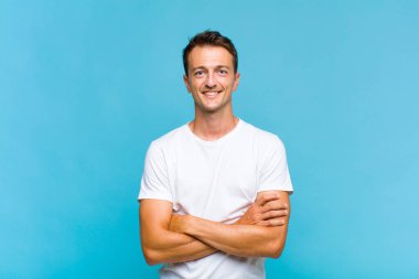 Genç yakışıklı adam kameraya çapraz kollarıyla gülümsüyor ve mutlu, kendinden emin, tatmin olmuş bir ifade, yan bakış.