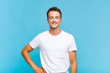 Genç yakışıklı adam mutlu bir şekilde gülümsüyor, eli kalçasında ve kendinden emin, olumlu, gururlu ve arkadaş canlısı.