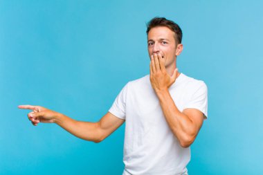 Genç yakışıklı adam kendini mutlu, şaşkın ve şaşırmış hissediyor. Ağzını eliyle kapatıyor ve yanal kopya alanını işaret ediyor.