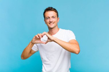 Genç yakışıklı adam gülümsüyor ve mutlu, sevimli, romantik ve aşık, iki eliyle kalbi şekillendiriyor.