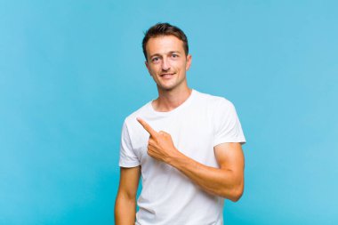 Genç yakışıklı adam neşeyle gülümsüyor, mutlu hissediyor ve kenarı ve yukarıyı işaret ediyor, fotokopi uzayındaki nesneleri gösteriyor.