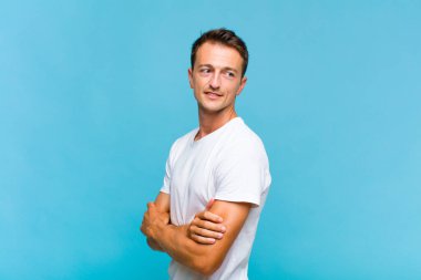 Genç yakışıklı adam neşeyle gülümsüyor, mutlu, tatmin olmuş ve rahatlamış hissediyor, kollarını kavuşturup yan tarafa bakıyor.