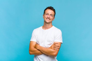 Genç yakışıklı adam kollarını kavuşturup mutlu bir şekilde gülüyor, rahat, pozitif ve tatmin edici bir pozla.