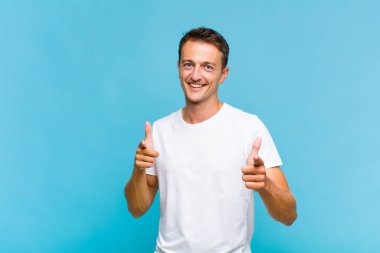 Olumlu, başarılı, mutlu bir tavırla gülümseyen genç yakışıklı adam kamerayı işaret ediyor, elleriyle silah işareti yapıyor.