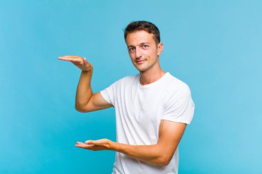 Genç yakışıklı bir adam iki eli de yan tarafında fotokopi alanı olan bir nesneyi tutuyor, bir nesneyi gösteriyor, sunuyor ya da reklamını yapıyor.