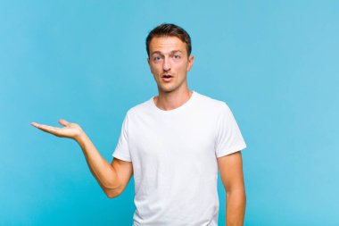 Genç yakışıklı adam şaşırmış ve şoka uğramış görünüyor, çenesi düşmüş, eli açık bir nesneyi tutuyor.