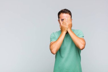 Genç yakışıklı adam elleriyle yüzünü kapatıyor, parmaklarının arasından şaşkın bir ifadeyle yan tarafa bakıyor.