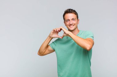 Genç yakışıklı adam gülümsüyor ve mutlu, sevimli, romantik ve aşık, iki eliyle kalbi şekillendiriyor.