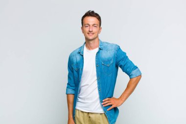 Genç yakışıklı adam mutlu bir şekilde gülümsüyor, eli kalçasında ve kendinden emin, olumlu, gururlu ve arkadaş canlısı.