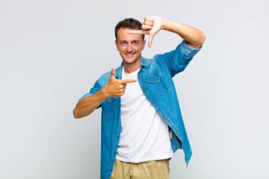 Genç yakışıklı adam mutlu, arkadaş canlısı ve pozitif hissediyor, gülümsüyor ve elleriyle bir portre veya fotoğraf çerçevesi yapıyor.