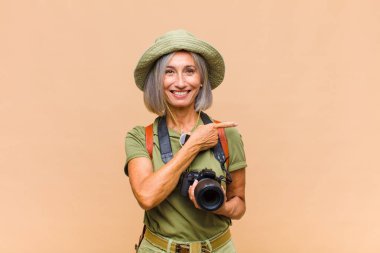 Orta yaşlı kadın neşeli bir şekilde gülümsüyor, mutlu hissediyor ve yan ve yukarı doğru işaret ediyor, fotokopi uzayındaki nesneleri gösteriyor.