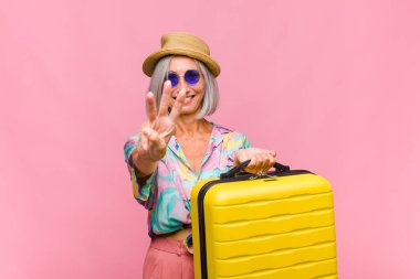 Orta yaşlı bir kadın gülümsüyor ve arkadaş canlısı görünüyor. Üç ya da üç numarayı gösteriyor. Elleri önde, geri sayıyor.