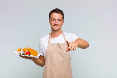 Genç ve yakışıklı bir adam kameraya bakıyor. Tatmin olmuş, kendine güvenen, dost canlısı bir gülümsemeyle seni seçiyor.
