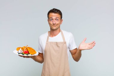 Genç yakışıklı adam şaşkın ve şaşkın, şüpheci, farklı seçenekleri tartıyor ya da komik bir ifadeyle seçiyor.