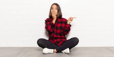 İspanyol kadın kendini mutlu, şaşkın ve şaşırmış hissediyor. Ağzını eliyle kapatıyor ve yanal kopya alanını işaret ediyor.