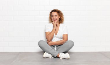 Orta yaşlı bir kadın mutlu, kendinden emin bir ifadeyle gülümsüyor, elleri çenesinde, merak ediyor ve yan tarafa bakıyor.