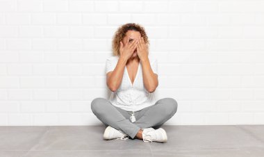 Orta yaşlı kadın elleriyle yüzünü kapatıyor, parmaklarının arasından şaşkın bir ifadeyle yan tarafa bakıyor.