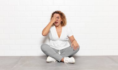 Orta yaşlı bir kadın şok olmuş, korkmuş ya da korkmuş görünüyor. Yüzü elle kaplanmış ve parmaklarının arasından gizlice bakıyor.