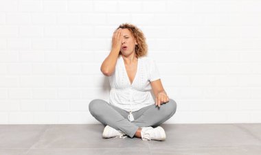 Orta yaşlı bir kadın uykulu, sıkılmış ve esniyor, baş ağrısı ve yüzünün yarısını kaplayan bir elle.