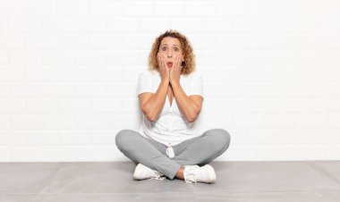 Orta yaşlı kadın şok olmuş ve korkmuş hissediyor, ağzı açık ve elleri yanaklarında korkmuş görünüyor.