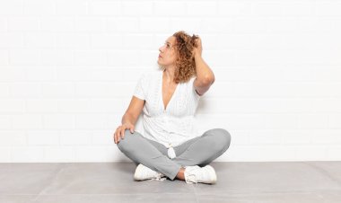 Orta yaşlı bir kadın bilgisiz ve kafası karışık, bir çözüm düşünüyor, eli kalçasında, diğeri kafasında, dikiz aynasından.