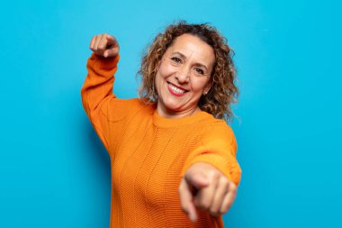 Orta yaşlı bir kadın mutlu ve kendinden emin, kamerayı iki eliyle gösteriyor ve gülüyor, seni seçiyor.