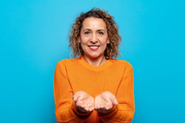 Orta yaşlı kadın mutlu bir şekilde gülümsüyor arkadaş canlısı, kendinden emin, olumlu bakışlı, bir nesne ya da konsept sunuyor ve gösteriyor.