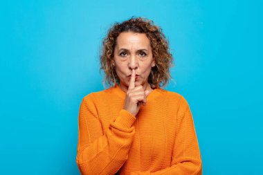 Orta yaşlı kadın ciddi görünüyor ve parmaklarını dudaklarına bastırıp sessizlik ya da sessizlik talep ediyor, sır saklıyor.